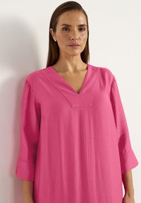Rosa Kurzarmkleid mit V-Ausschnitt und lockerer Passform. Weicher, glatter Stoff mit leicht strukturierten Oberfläche. Mit Ellenbogenlangen Ärmeln.