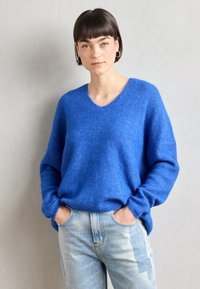 Blue knitted sweater with a V-neck and loose fit, featuring long sleeves and ribbed texture. Paired with light blue jeans.