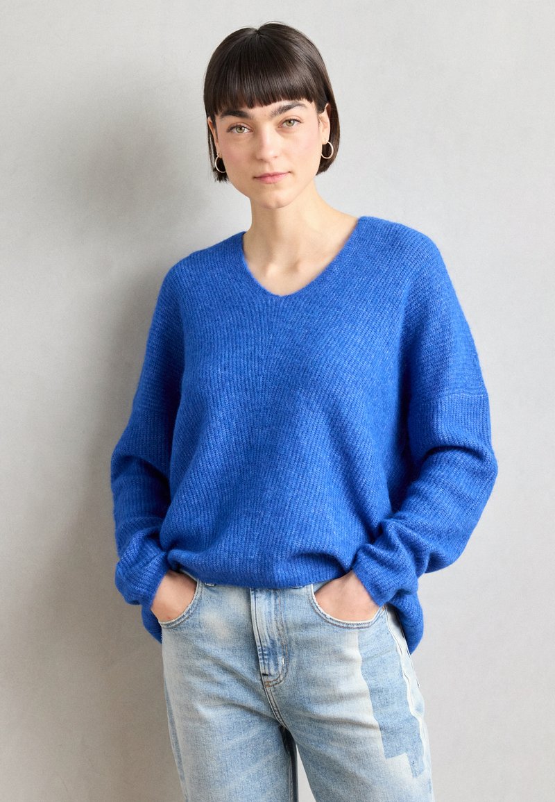 Blue knitted sweater with a V-neck and loose fit, featuring long sleeves and ribbed texture. Paired with light blue jeans.