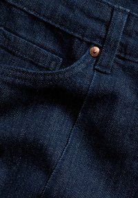 Dunkelblauer Denim-Stoff im Close-up mit einem kupferfarbenen Knopf, der eine strukturierte Oberfläche, kontrastierende Nähte und eine glatte Textur aufweist.