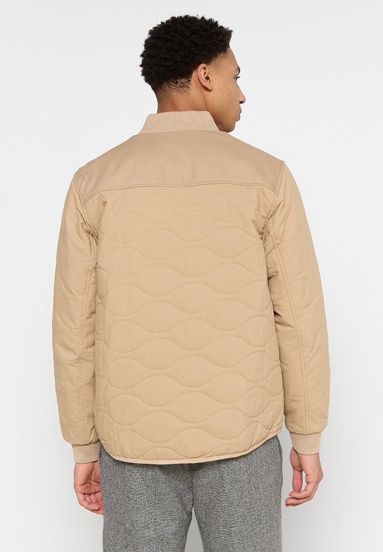 Giacca trapuntata beige con collo a coste e motivo testurizzato, caratterizzata da una linea dritta e maniche lunghe, indossata sopra pantaloni grigi.