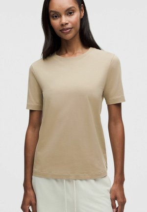 Mujer con camiseta lisa de manga corta beige y pantalones de color claro sobre un fondo neutral, mirando hacia adelante con expresión neutra.