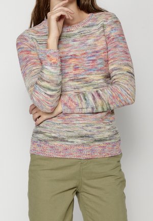 Personne portant un pull tricoté multicolore aux manches longues et un pantalon vert olive, les bras croisés et la main touchant le visage.