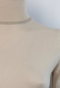 Maglione a collo alto beige con una texture a costine, caratterizzato da un bordo scalloppato al colletto. Realizzato in materiale morbido, presenta un design senza cuciture.