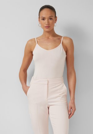 Kobieta z włosami zaczesanymi do tyłu, nosząca kolczyki koła, jasnoróżowy top bez rękawów oraz dopasowane spodnie z wysokim stanem, stoi na tle szarego tła.