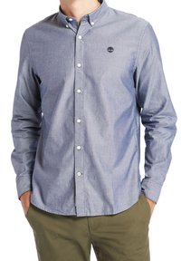 Uomo che indossa una camicia azzurra chiara con il logo Timberland sul petto e pantaloni verde oliva, con le mani nelle tasche, in piedi contro uno sfondo bianco.