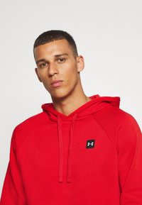 Rode hoodie met een kangoeroezak, platte trekkoorden en een klein zwart logo-opdruk op de borst. Zachte, gestructureerde stof.