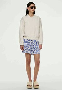 Veste bomber beige clair, shorts bleu et blanc imprimés, et sandales à plateforme beiges, accompagnés de lunettes de soleil surdimensionnées.