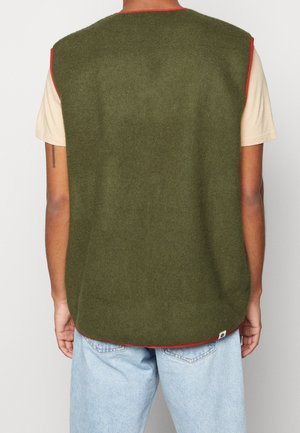 Rückansicht einer Person, die eine olivgrüne ärmellose Weste mit rotem Rand über einem beige-farbenen Kurzarmhemd und hellblauen Jeans trägt.