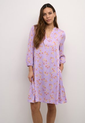 Femme aux longs cheveux bruns portant une robe violette à fleurs, midi et à manches 3/4, sur un fond clair uni.