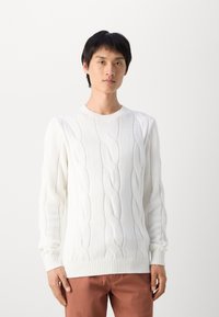 Nuori mies, jolla on lyhyet mustat hiukset, pukeutuneena valkoiseen pajupaitaan ja ruosteenvärisiin housuihin, seisoo tasaisen vaaleataustan edessä.