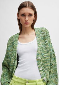 Grønn strikket cardigan med et teksturert mønster, som har en avslappet passform, knapper foran, og er kombinert med en hvit tank topp og grønne bukser.