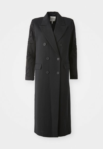 Melnā divrindas mētelī ar asu apkakli, sešiem pogām un pērlītēm uz piedurknēm. Gluda auduma, izpildīts pielāgotā formā un garā garumā.