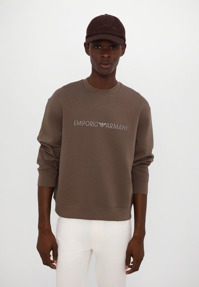 Brown sweatshirt with embroidered "Emporio Armani" logo, round neck, long sleeves. Worn with white pants and a brown cap.