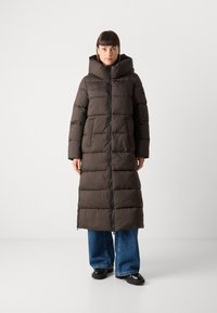 Lange pufferjas in donkerbruin, met een hoge kraag, twee voorzakken en een ritsluiting. Horizontaal gestikte patroon en oversized vorm.
