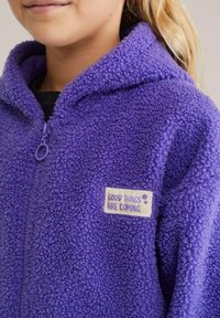 Violets fleece sporta jakas ar teksturētu, pūkainu virsmu. Iekļauta rāvējslēdzēja aizdare un neliels uzšuves gabals ar tekstu "LABAS LIETAS NĀK".