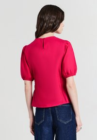Donna con capelli scuri e mossi che indossa una blusa a maniche corte di un brillante rosa, con maniche a palloncino, e jeans blu scuro, vista di spalle.