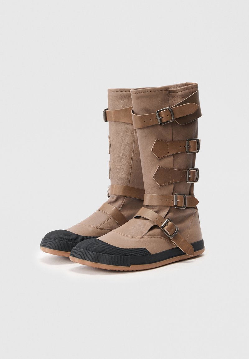 Tan high-top boots made of leather and fabric, featuring multiple adjustable straps and a black rubber toe cap for added durability.