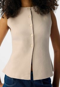 Ärm-lös beige knapplöst linne med en lätt flare, bärs med mörkblå jeans som syns vid midjan.