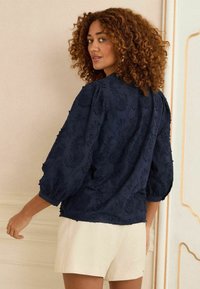 Blusa ricamata blu navy con maniche a pallone, motivi floreali testurizzati e scollatura rotonda, abbinata a pantaloni corti di colore chiaro.