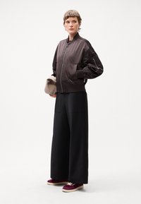 Brown bomber jacket with ribbed cuffs, paired with wide leg black trousers and burgundy sneakers. Model holds a beige clutch bag.