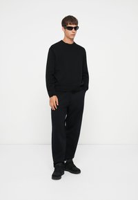Schwarzer Strickpullover mit geripptem Saum, kombiniert mit lässigen schwarzen Sweatpants. Das Modell trägt schwarze Sonnenbrillen und schwarze Sneakers. Minimalistisches Design.