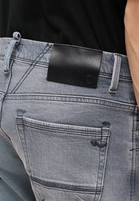 Πίσω τσέπη τζιν γαλάζιου χρώματος με κεντημένο λογότυπο και μαύρη δερμάτινη ετικέτα μάρκας στη ζώνη, φορεμένο με μαύρο πουκάμισο.