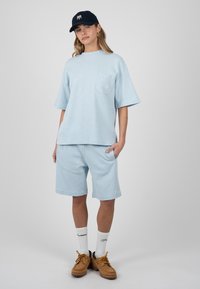 Femme debout portant un T-shirt oversize bleu clair et un short, casquette marine, chaussettes blanches et chaussures marron à lacets sur fond uni.