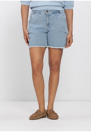 Vrouw draagt lichtblauwe spijkershorts met rafelige zomen, een lichtblauwe trui en bruine instapsloffen, staand tegen een effen achtergrond.