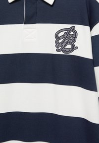 Polo majica s plavim i bijelim prugama, izrađena od teksturirane tkanine, s ovratnikom na dugme i izvezenim logotipom na lijevoj strani prsiju.