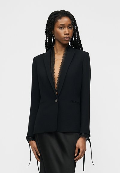 Femme aux longues tresses portant un blazer noir ajusté avec une bordure en dentelle et une jupe en satin noir assortie sur un fond clair uni.