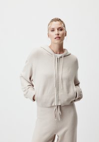 Felpa beige con zip, dotata di polsini e orlo a coste, realizzata in materiale morbido. Presenta un cappuccio con cordino e un discreto dettaglio del logo sul petto.