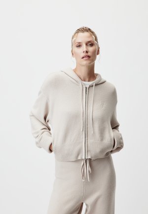 Béžová mikina na zips s rebrovanými manžetami a spodným lemom, vyrobená z mäkkého materiálu. Obsahuje kapucňu so šnúrkou a nenápadný logotyp na hrudníku.