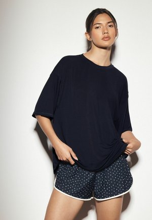 Ung kvinde iført en løs sort t-shirt og sorte shorts med hvide prikker, stående foran en ensartet lys baggrund.