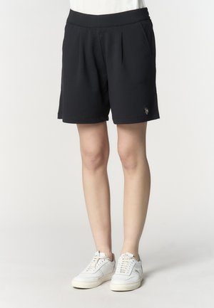 Persona che indossa pantaloncini neri plissettati e sneaker bianche, in piedi contro uno sfondo chiaro e uniforme.