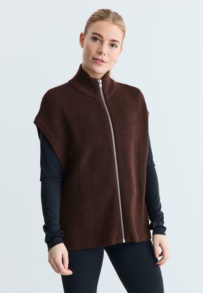 Bruine mouwloze gebreide vest met hoge kraag en voorrits, met ribbeltextuur en een losse pasvorm. Draag over een zwart langemouwen shirt.