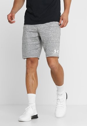Mężczyzna ubrany w szare spodenki Under Armour, białe skarpetki sportowe i białe buty sportowe, stojący na jednolitym jasnym tle.