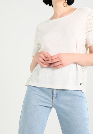 White linen t-shirt with short sleeves featuring lace detailing. Paired with light blue denim jeans. Simple cut and relaxed fit design.