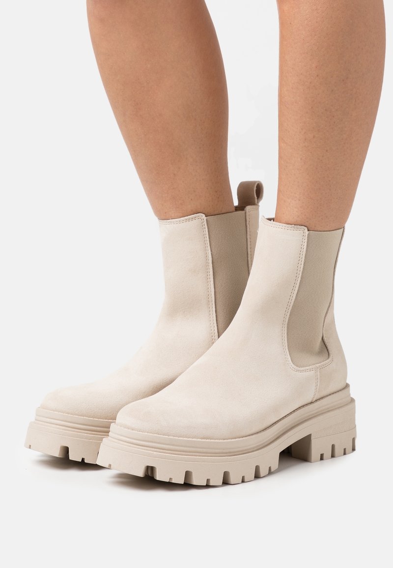 Bottes cheville en daim beige avec semelles épaisses à crampons et panneaux latéraux élastiques, portées sur jambes nues sur un fond uni.