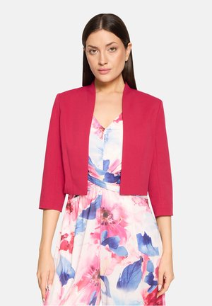 Woman with long dark hair wearing a bright pink cropped blazer over a floral dress with pink, blue, and white colors on a plain background.