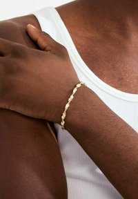 Kuldne käevõru keerulise mustriga, millel on tekstureeritud lingid, kantud tumeda nahaga käsivarrel. Taustal valge soonikkoes tanktop.