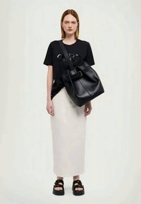 Μαύρη δερμάτινη τσάντα κάδου με δομημένο σχήμα, καπάκι που διπλώνει και λεπτομέρεια από μεταλλικό κούμπωμα. Το μοντέλο φοράει μαύρο t-shirt, λευκή φούστα και χοντρές σαγιονάρες.