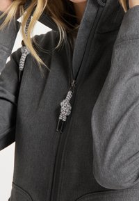 Närbild av en person som bär en mörkgrå zip-up hoodie med mönstrade snören, som håller huvan nära sitt huvud.