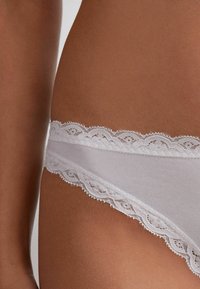 String en dentelle blanche avec une fine matière en coton. La dentelle a une bordure festonnée et est superposée pour un détail et une texture supplémentaires.