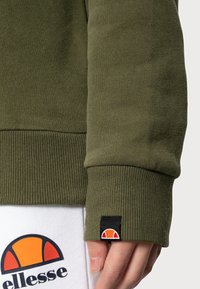 Felpa verde oliva con polsini a costine e una piccola etichetta con logo. Pantaloni felpati bianchi con un design grafico dai toni arancioni e neri.