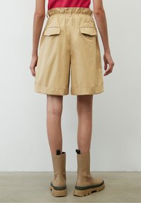 Persona che indossa pantaloni corti beige a vita alta con tasche posteriori e stivali beige chunky, in piedi contro una parete bianca semplice.