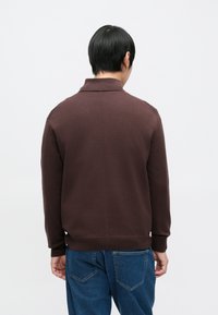 Brun strikket genser med en oppbrettet krage, lange ermer og ribbet nederkant. Har en glatt tekstur og minimalistisk design.