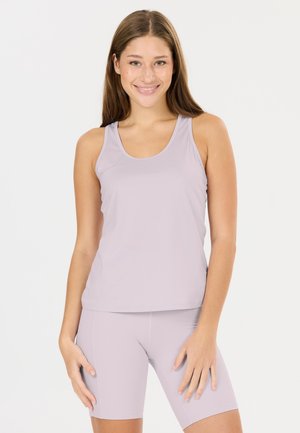 Jeune femme souriante, portant un débardeur sans manches violet clair et un short assorti ajusté, debout devant un fond blanc.