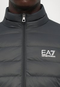 Nærbilde av en svart quilted jakke med glidelås foran og hvit EA7 Emporio Armani-logo på brystet.