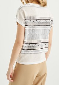 Donna che indossa un top bianco a maniche corte in maglia con motivi geometrici neri, abbinato a pantaloni beige, vista da dietro.
